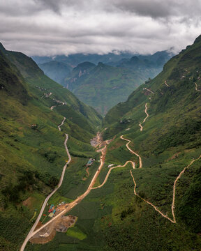 Aerial view of the winding mountain roads of Ha Giang, featuring lush green slopes, deep valleys, and dramatic cloud-covered peaks in Tien Du, Bac Ninh, Vietnam.