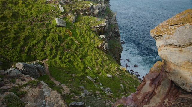 Aerial view of rugged coastal cliffs beside blue sea, with green vegetation, layered rock and waves below. Dramatic shoreline landscape highlights erosion, wild terrain and ocean scenery.