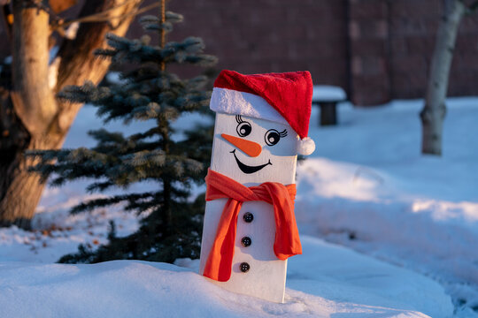 Cute decorative snowman made of painted white wooden plank wearing Santa hat and orange scarf standing in snow outside with evergreen pine tree background at sunset. Handmade Christmas decoration