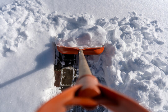 Snow removal with orange shovel, closeup. First person view of clearing fresh thick snow from brick pavement using bright orange plastic shovel with wooden handle on bright sunny winter day