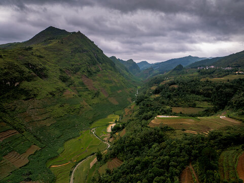 Aerial view of Ha Giang Loop featuring lush green mountains, terraced rice fields, and a winding stream under a cloudy sky, Dong Van, Tuyen Quang, Vietnam.