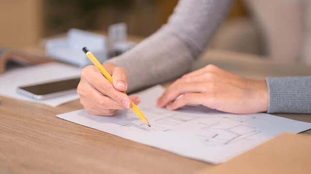 Female designer sketching furniture blueprint with yellow pencil on wooden workspace with coffee and reference materials