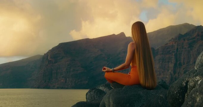 Woman meditating in lotus position on volcanic shore overlooking mountains