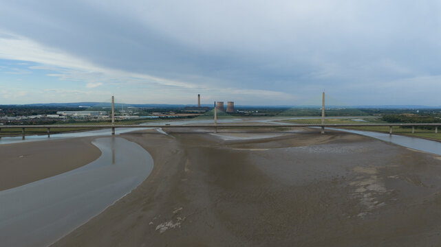 Aerial view of the Mersey Gateway Bridge spanning the River Mersey at low tide with industrial cooling towers in the distance under a cloudy sky in Runcorn, United Kingdom.