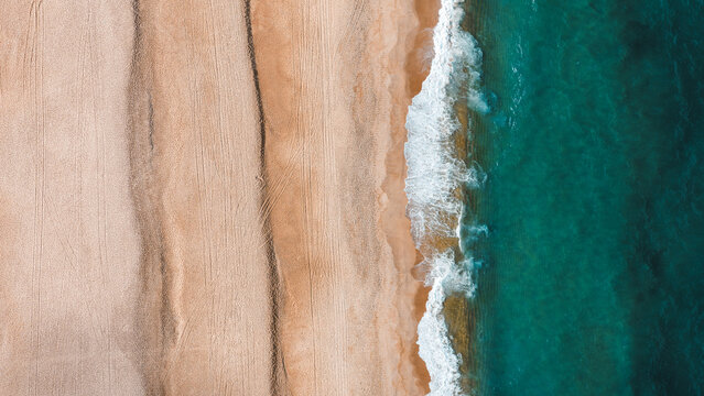 Aerial view of the sandy beach at Nazare with visible tire tracks and turquoise ocean waves crashing on the shore in Nazare, Leiria District, Portugal.