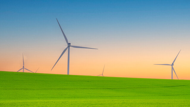Wind turbines rise above a vibrant green field at sunset, representing renewable energy, clean electricity, sustainability, green technology, climate action, and a low carbon future.