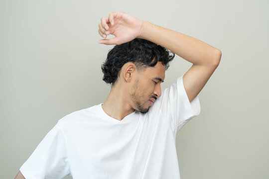 Young Asian man smelling his armpit feeling bad and worried about body odor or sweat problem wearing white t shirt standing over gray background male personal hygiene and health care concept