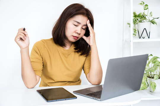 tired Asian business woman suffering from headache and fatigue while working on laptop and tablet at desk. Stressed female worker feeling burnout syndrome, overwhelmed, and exhausted in modern office