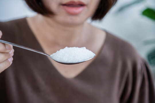 Close-up of Asian woman holding spoon of white refined sugar. Female eating excess sugar leading to sugar addiction, diabetes risk, unhealthy diet, obesity, and high blood glucose level