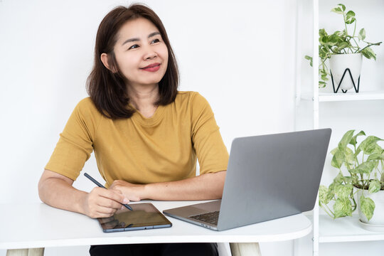 Happy Asian business woman smiling and thinking while working on laptop and digital tablet at desk. Cheerful female graphic designer, freelancer, or remote worker inspired in bright modern office