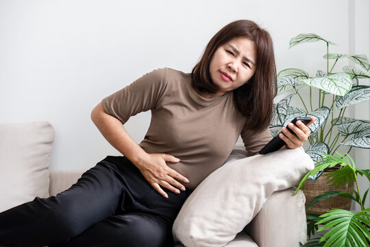 Asian woman lying on sofa suffering from severe stomachache, abdominal pain, and menstrual cramps . Sick female feeling indigestion, diarrhea, or illness at home