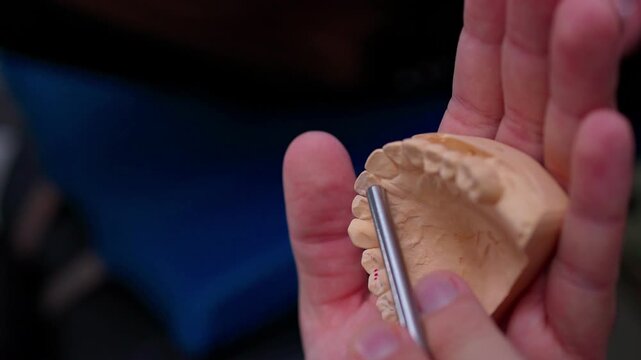 Hands probing upper dental plaster model with metal instrument during closeup instructional inspection, showing careful rotation, precise contact, and clinical laboratory work