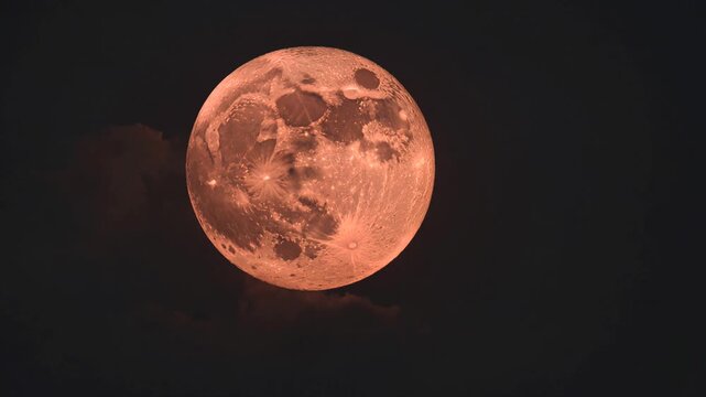 Huge red full moon rising in the night sky with dark mysterious clouds passing, revealing the detailed surface craters of our natural satellite in an amazing close up