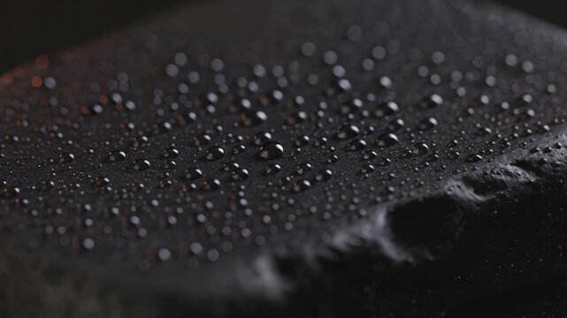 Water droplets glistening on a dark, textured black stone surface with warm light reflections, representing relaxation, spa therapy, and natural mineral purity in a close-up view