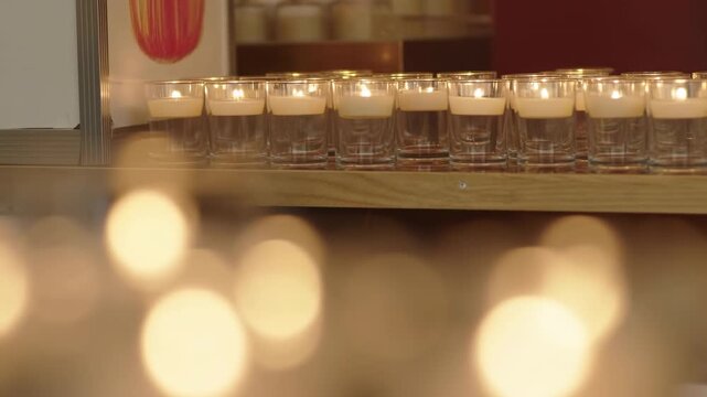 Warm glass candle holders on wooden shelf with soft golden bokeh lights, blurred foreground glow, cozy ambient interior mood, elegant decorative lighting and gentle amber atmosphere
