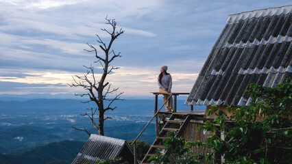 Fototapeta premium A woman sitting on wooden balcony, looking at mountain views on cloudy morning