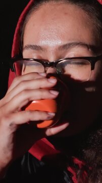 Female figure dons crimson hoodie as she repeatedly lifts. Cup remains close to face as subject moves hand. Controlled light creates contrast between bright orange object