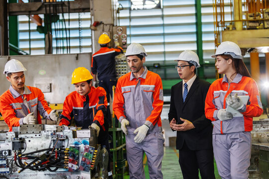 Factory manager and engineering team inspect machinery and production work together on a manufacturing floor. Skilled workers examine equipment while management observes the process,