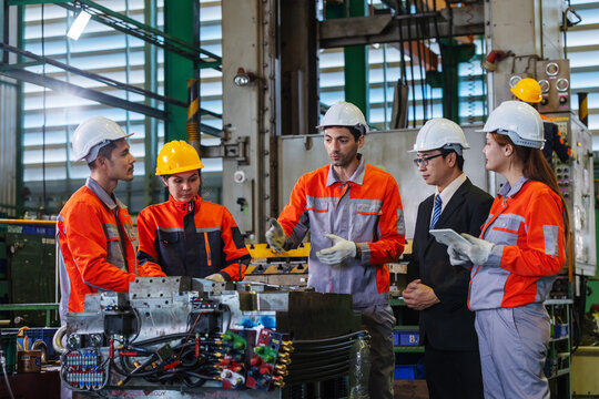 Factory workers, engineers and manager hold a team meeting beside industrial machinery to discuss work instructions and production processes.