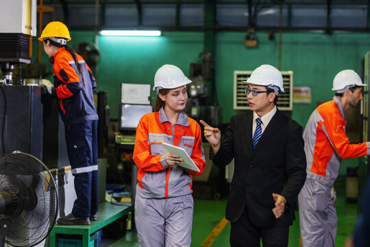 Factory manager and female supervisor discuss production while walking through a busy manufacturing facility. Workers operate machinery in the background as management reviews factory operations