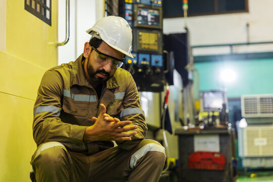 Tired industrial worker wearing safety equipment sits and rests beside machinery during a break in a manufacturing factory.