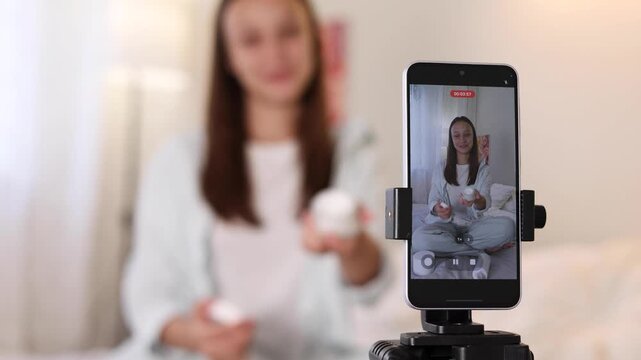 Teenage girl filming beauty blog at home while reviewing skincare product for social media audience. 