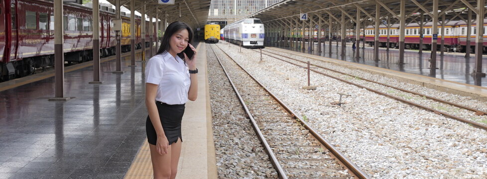 A young Asian woman with long black hair in a university uniform stands on a train station platform while holding a phone to her ear