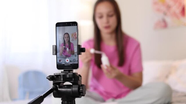 Teenage girl filming beauty blog at home while reviewing skincare product for social media audience. 