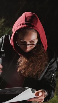 Young woman in red hooded sweatshirt examines open book. Glasses frame remains clear on face near open book. Consistent close up framing captures details of hair clothing