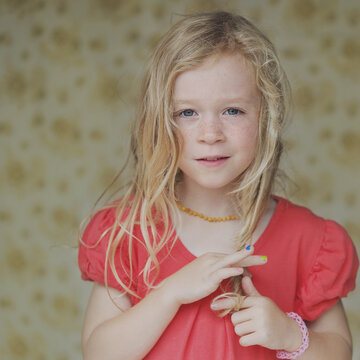 Portrait of a girl with long blonde hair standing in front of a wallpaper covered wall playing with her hair