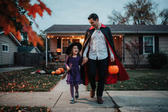 Father in vampire cape walking with young daughter on suburban sidewalk at dusk, autumn leaves and cozy house lights, Halloween family costume