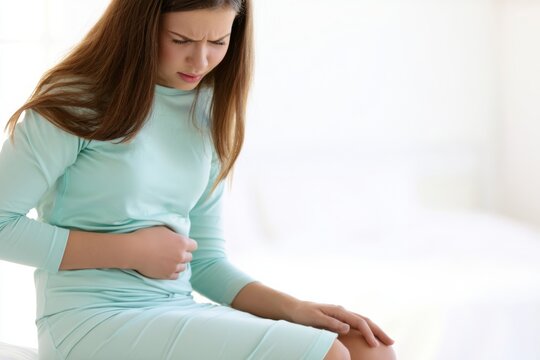 Young woman in light blue dress sitting on bed, experiencing menstrual cramps with one hand on her abdomen and a tense posture in a softly lit room