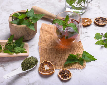 Fresh nettle leaves and dried herbs with a glass cup of herbal tea, dried lemon slices, mortar and burlap on a marble surface