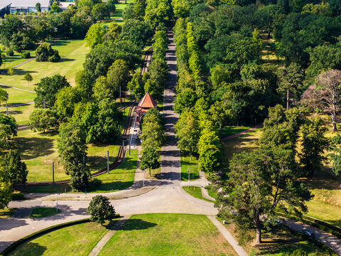 Aerial view of Herrenkrug in Magdeburg shows tree-lined avenue beside tram tracks, park lawns and station shelter. Summer landscape combines public transport, greenery and quiet urban recreation