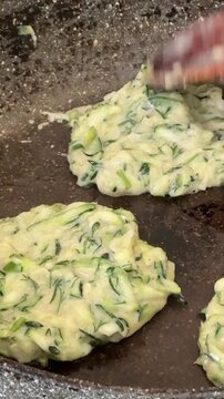Zucchini pancakes frying in a hot skillet. Overhead close-up shot of a wooden spoon flattening savory vegetable fritters in a non-stick pan.