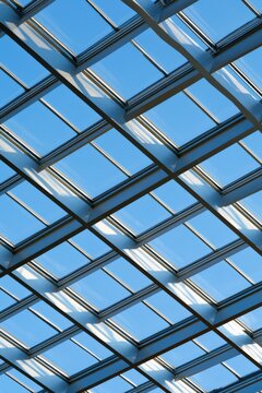 Modern architectural glass roof viewed from below, featuring a geometric grid of metallic beams and large blue-tinted glass panels against a clear sky