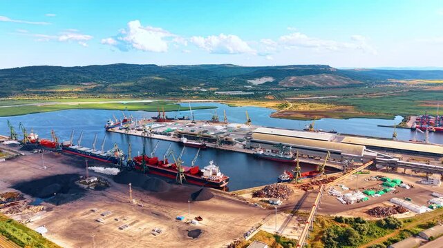 Cargo seaport terminal with docked ships and harbor cranes. High angle view of industrial port, scrap metal piles, blue water, green hills, clear sky.