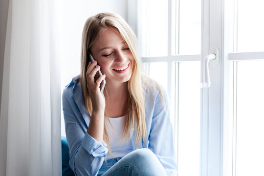 Happy woman talking on mobile phone at home by window. Dreamy girl holding cell phone and calling, enjoying conversation. People connection, communication. Lifestyle moment