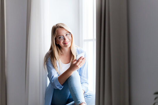 Young woman using smartphone, sitting on windowsill. Happy girl relaxing by window. Distance learning, online education. Freelancer working from home office. People connection, communication