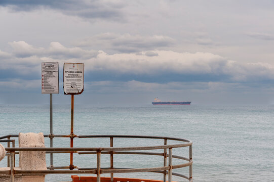 Cartelli di avviso sul lungomare con vista sul mare e nave all&rsquo;orizzonte sotto cielo nuvoloso. Scena costiera ideale per temi sicurezza, paesaggi marini, turismo e atmosfera fuori stagione.