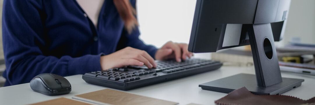 Young Asian furniture designer typing on computer while reviewing material samples and color swatches at modern studio desk