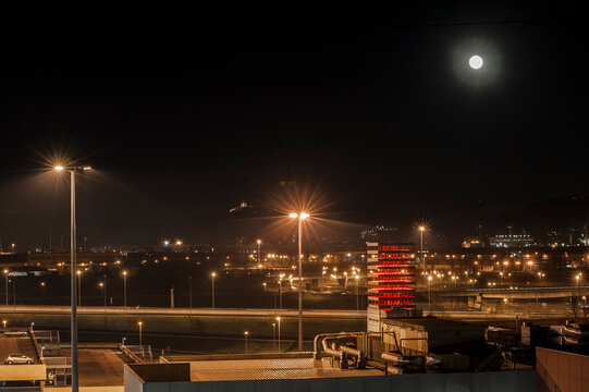 Veduta notturna con luna piena, luci urbane e strutture industriali illuminate. Ideale per temi cityscape, infrastrutture, energia, atmosfera notturna, paesaggio urbano e fotografia ambientale.