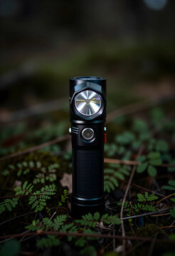 Black tactical flashlight standing upright on a forest floor with green foliage and dark background, the flashlight features a textured grip and a prominent power button