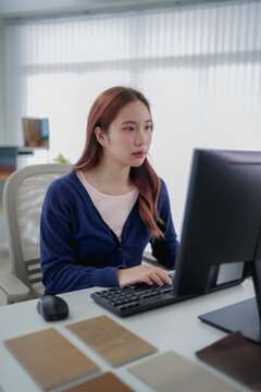 Young Asian furniture designer reviewing material samples while working on computer in modern design office