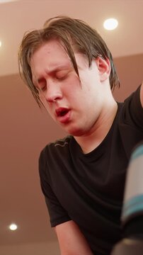 Close up of young man with wet hair wearing black shirt indoors. Man holds and flexes his arm in boxing gloves under warm ceiling. Focus is on upper body movements