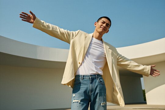 Young asian man opens arms wide outdoors. He wears light jacket white t-shirt ripped jeans. Bright sunny sky forms backdrop. Man looks happy.
