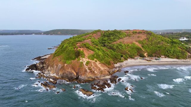 Cinematic aerial drone view of Vagator Beach cliffs at sunset, Goa, India. Tropical coastline meets ocean waves with red rocks and golden hour sky. Perfect for travel and tourism.