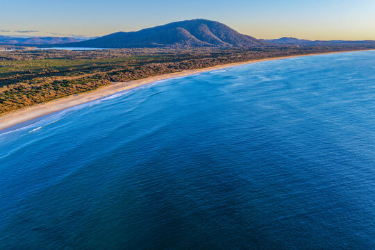 Aerial view along the long sandy shoreline of Diamond Head Beach, with the Pacific Ocean stretching beside the coast and a scenic mountain landscape rising in the background at Crowdy Bay 