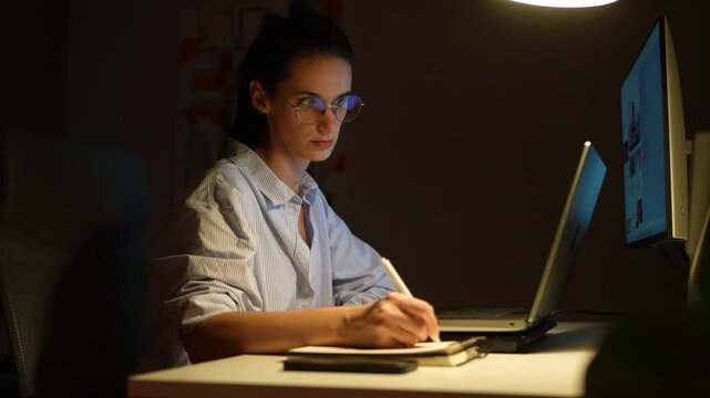 Businesswoman working late from home office writing notes and working on laptop