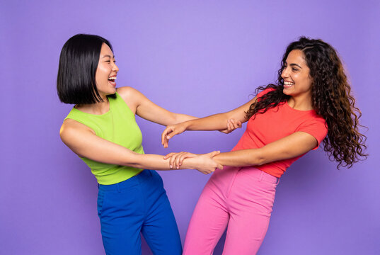 Two young female best friends laughing and playfully pulling each other by the hands against a vibrant lavender studio background.